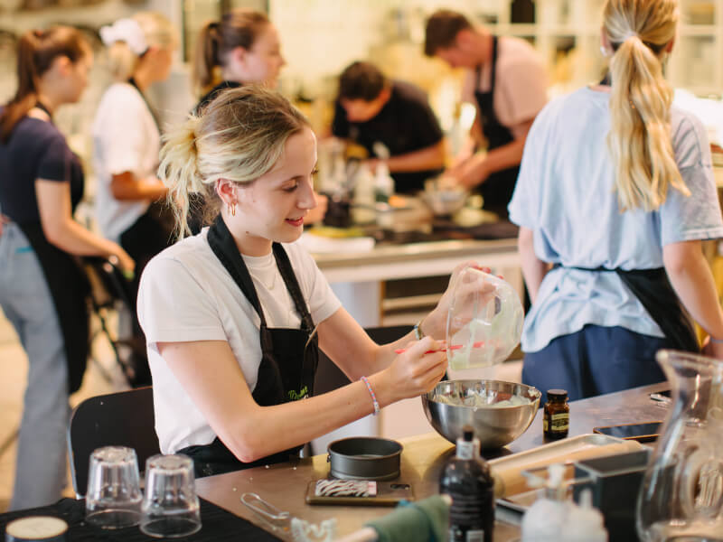 A woman sits at a table at a cooking class, pouring a mixture from one bowl to another